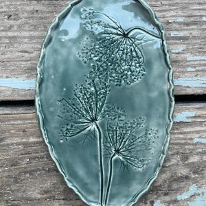 Könnte beinhalten: Ovale, türkisfarbene Keramikplatte mit strukturiertem, gewelltem Rand. Die Platte zeigt ein erhabenes, detailliertes botanisches Design einer blühenden Pflanze. Das Design ist auf der Platte zentriert, mit den Stielen und Blütenköpfen der Pflanze deutlich sichtbar.