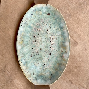 May include: Oval ceramic serving platter with a turquoise and speckled glaze. The platter has a brown rim and two brown decorative crown-shaped handles. The handmade dish is suitable for serving appetizers or displaying decorative items.