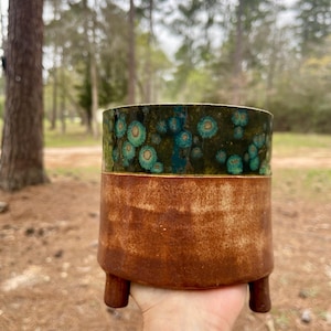 May include: A handcrafted ceramic planter with a two-tone design. The top section features a dark green glaze with turquoise accents, while the lower portion has a rustic brown finish. The planter is elevated by three small, rounded legs.