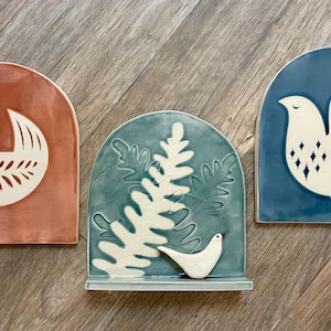 May include: Three arched ceramic wall plaques with glossy finishes. The left plaque is terracotta with a white bird design. The center plaque is teal with a white fern and bird. The right plaque is dark blue with a white bird featuring diamond cutouts.