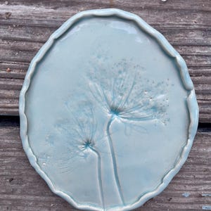 Puede incluir: Un plato de cerámica azul claro con borde festoneado. El plato presenta un delicado diseño en relieve de una planta con dos cabezas de flores. El plato es redondo y parece hecho a mano.