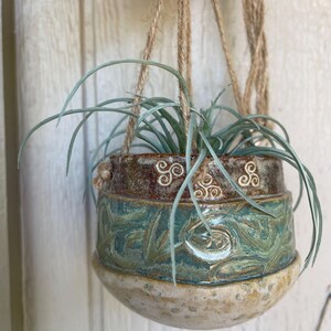 May include: A hanging ceramic planter with a plant. The planter is a mix of brown, teal, and beige with embossed Celtic knot designs. The plant has long, thin, light green leaves. The planter hangs from jute rope.