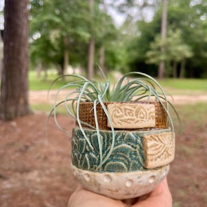 Pode incluir: Um pequeno vaso de cerâmica redondo com três faixas horizontais distintas. A faixa superior é castanha com textura de grelha, a do meio é azul-petróleo com relevo floral e a base é branca salpicada com pequenas covinhas.