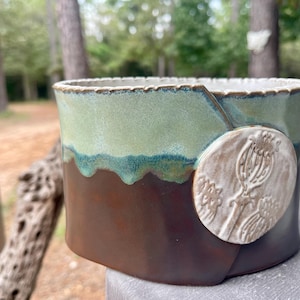 May include: A cylindrical ceramic yarn bowl featuring a two-tone glaze with a sage green top and a dark brown base. A circular white clay medallion is attached to the side, embossed with a detailed botanical illustration of three poppy seed pods.