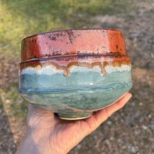 Könnte beinhalten: Eine handgefertigte Keramikschale mit einem einzigartigen Design. Der obere Bereich ist rostorange, geht in ein hellbraunes Band über und dann in einen türkisgrünen Sockel. Die Schale hat eine leicht unregelmäßige Form, was ihren handwerklichen Charme unterstreicht.