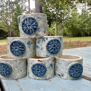 May include: A set of six cylindrical ceramic napkin rings with a speckled light blue and white glaze. Each ring features a raised, circular dark blue medallion with a detailed, embossed snowflake or floral pattern centered on the front surface.