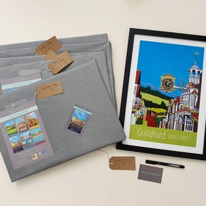 May include: A framed print of a colorful illustration of a street scene in Guildford, England. The print is titled "Guildford High Street". The print is surrounded by gray wrapping paper and a black marker pen. There are several tags with handwritten text on them.