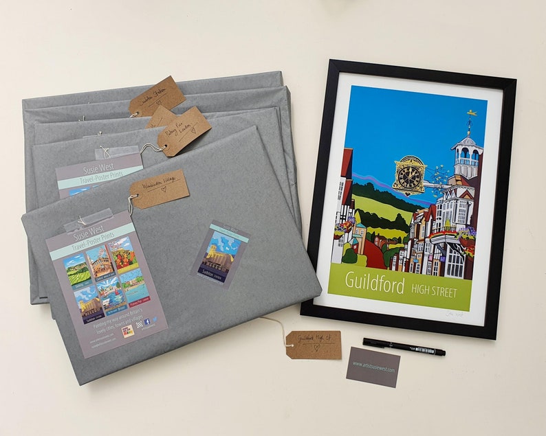 May include: A framed print of a colorful illustration of a street scene in Guildford, England. The print is titled "Guildford High Street". The print is surrounded by gray wrapping paper and a black marker pen. There are several tags with handwritten text on them.