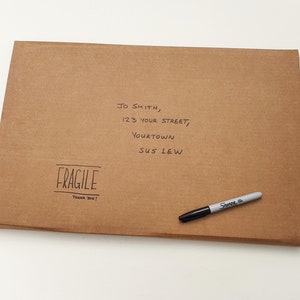 May include: A brown cardboard box with a handwritten address label and a "FRAGILE" sticker. The address reads "JO SMITH, 123 YOUR STREET, YOURTOWN SUS LEW". A black marker pen is lying on top of the box.