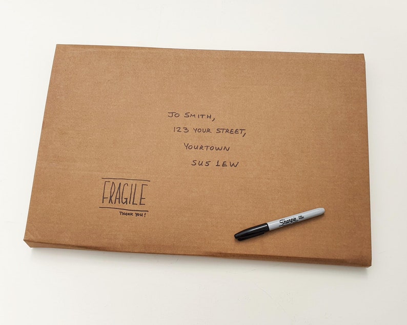 May include: A brown cardboard box with a handwritten address label and a "FRAGILE" sticker. The address reads "JO SMITH, 123 YOUR STREET, YOURTOWN SUS LEW". A black marker pen is lying on top of the box.