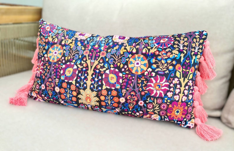 May include: A long rectangular pillow with a black background and a colorful floral pattern. The pillow has pink tassels on each end.