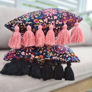 May include: Two decorative throw pillows with black and white floral patterns and tassels. The top pillow has pink tassels and the bottom pillow has black tassels.