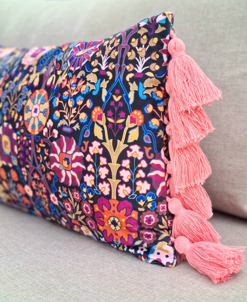 May include: A decorative pillow with a black background and a colorful floral pattern. The pillow has pink tassels on the edges.
