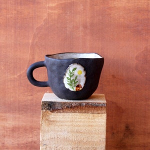 Puede incluir: Una taza de cerámica negra hecha a mano con una forma irregular única. La taza presenta un diseño ovalado pintado con una casa, un sol y follaje. El interior es blanco. La taza está sobre un bloque de madera sobre un fondo marrón.