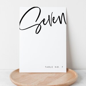May include: White table number card with black calligraphy that says "Seven" and the text "Table No. 7" at the bottom.