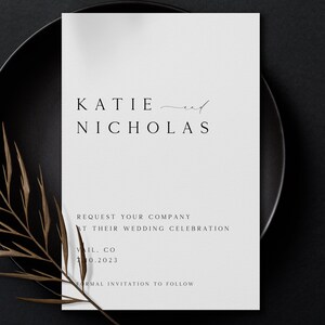 May include: A white wedding announcement card with black text. The card reads "KATIE and NICHOLAS REQUEST YOUR COMPANY AT THEIR WEDDING CELEBRATION VIL. CO 10.2023 FORMAL INVITATION TO FOLLOW."