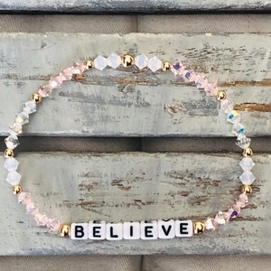 May include: A delicate bracelet featuring the word "BELIEVE" spelled out in white letter beads. The bracelet is adorned with small, iridescent crystals and pink and clear beads, interspersed with gold-colored beads.