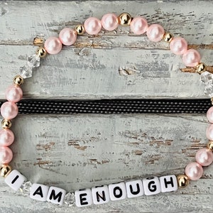May include: A pink and gold beaded bracelet with clear crystal beads and white letter beads spelling "I AM ENOUGH".