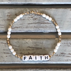 May include: White and gold beaded bracelet with the word "FAITH" spelled out in white letter beads.