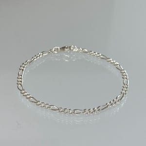 May include: A silver Figaro chain bracelet. The bracelet has an alternating pattern of elongated and round links, fastened with a lobster clasp. The jewellery is shown on a reflective surface, emphasising its polished finish.