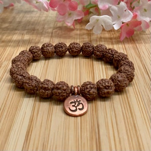 May include: A brown beaded bracelet with a copper Om charm. The bracelet is made of natural beads and has a simple, elegant design.