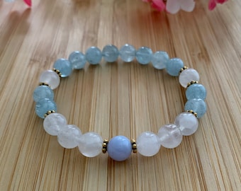 May include: A bracelet made with light blue aquamarine beads, white quartz beads, and a single light blue agate bead. The bracelet is on a wooden surface.