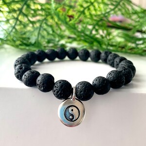 May include: Black lava rock bead bracelet with a silver Yin Yang charm. The bracelet has round, porous beads. The charm is a circular pendant with the Yin Yang symbol.