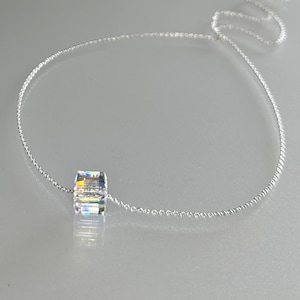 May include: A delicate silver necklace featuring a clear, iridescent cube pendant. The pendant reflects multiple colors. The chain is thin and appears to be made of small, interconnected links. The necklace is displayed on a light gray surface.