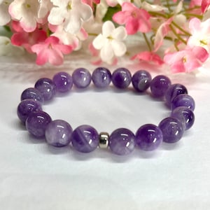 Puede incluir: Un brazalete de amatista púrpura con un cierre plateado. El brazalete está hecho de cuentas redondas y está enhebrado en elástico.