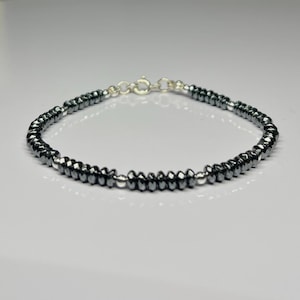 May include: A bracelet composed of small, faceted, dark grey metallic beads arranged in a circular pattern. The piece features interspersed smooth, polished silver-toned spherical beads and is secured with a silver-toned metal spring ring clasp.
