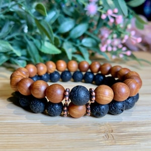 May include: Two bracelets, one made of brown wood beads and the other of black lava rock beads, are stacked on top of each other. The bracelets are connected by copper spacer beads.