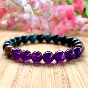 May include: A beaded bracelet featuring a mix of gemstones. The bracelet showcases a row of deep purple amethyst beads, transitioning to dark green and black beads. The bracelet is displayed on a wooden surface, with pink flowers in the background.