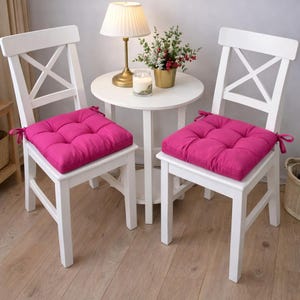 May include: Two white wooden chairs with magenta seat cushions. The cushions are square and tufted. A small white table with a lamp, candle, and flowers sits between the chairs. The chairs have an X-shaped back.