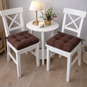 May include: Two white wooden chairs with brown seat cushions. The cushions are square-shaped and tufted. A small white table with a lamp and a floral arrangement sits between the chairs.