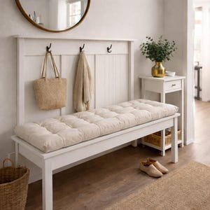 May include: A white wooden entryway bench with a tufted beige cushion. A matching white backboard features three black hooks, with a straw tote bag and a beige scarf hanging from them. A round mirror hangs above.