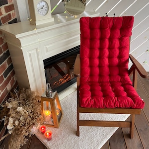 May include: A red padded chair with a wooden frame sits in front of a white fireplace. The chair has a tufted back and seat. The fireplace has a realistic flame effect.