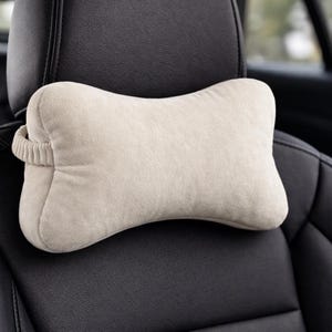 May include: A beige car headrest pillow with a bone-shaped design. The pillow is made of a soft, plush material and has a loop on the side for attachment. The headrest is positioned on a black car seat.