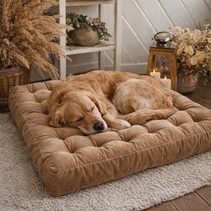 May include: A sleeping golden retriever rests on a tufted, square dog bed. The bed is a warm, muted brown color and sits on a light-colored rug. The dog's fur matches the bed's color, creating a cozy and inviting scene.