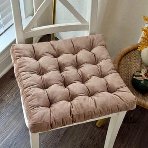 May include: A square, tufted seat cushion in a muted rose color rests on a white wooden chair. The cushion has a soft, velvety texture and is designed with a grid pattern of stitched squares. The chair is positioned near a window.
