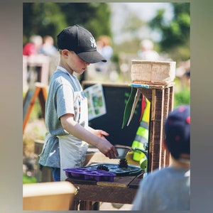 May include: A young person wearing a black baseball cap and a white apron is playing with a wooden play kitchen. The kitchen has a sink, a stove, and a counter. There are purple silicone moulds on the counter.