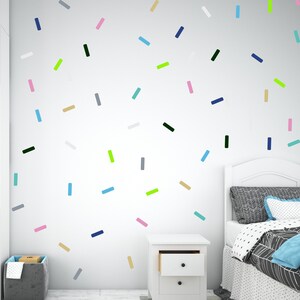 May include: A white wall decorated with colourful rectangular stickers in various colours, including pink, blue, green, black, and grey. The wall is decorated with a bed with a white headboard and a black and white patterned duvet. A white bedside table with two drawers is in the lower right corner of the image.