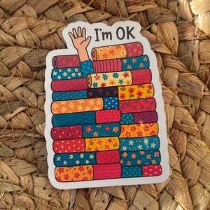 Può includere: Un adesivo con la scritta "I'm OK" presenta una pila di libri colorati e decorati. Una mano esce dai libri. I libri hanno disegni floreali e geometrici nei toni del rosso, blu, arancione e giallo.
