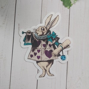 Pode incluir: Um coelho branco vestindo um colete roxo com cora&ccedil;&otilde;es e uma gravata borboleta azul, segurando um pergaminho e uma trombeta. O coelho &eacute; um adesivo com um toque de brilho azul.
