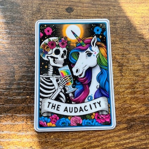 May include: A rectangular sticker featuring a skeleton and a unicorn. The skeleton wears a floral crown and holds a rainbow-colored drink. The unicorn has a rainbow mane and horn. The text "THE AUDACITY" is on a banner below.