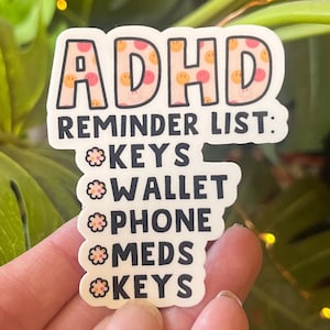 May include: A white sticker with a pink and orange background featuring a smiling face pattern. The sticker says "ADHD Reminder List:" and lists "Keys, Wallet, Phone, Meds, Keys".
