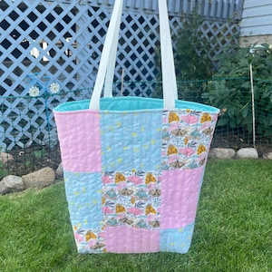 May include: A patchwork tote bag in shades of pink and light blue, with a floral pattern on some squares. The bag has a turquoise interior and white handles. The bag is designed with a quilted pattern.