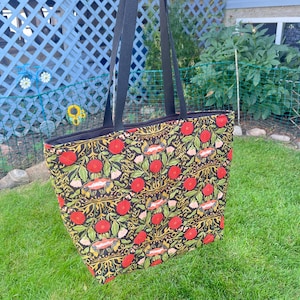 May include: A tote bag with black handles and a black zip closure. The bag displays a repeating pattern of red flowers, green leaves, and gold details on a black backdrop. The bag is held up against a backdrop of green grass and a blue fence.