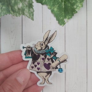 Pode incluir: Adesivo de coelho branco com um la&ccedil;o azul, segurando um pergaminho e uma trombeta. O coelho est&aacute; vestindo um colete roxo e branco com cora&ccedil;&otilde;es. O adesivo tem um detalhe de joia azul.