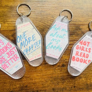 May include: Four clear acrylic keychains with silver key rings. Each keychain has a retro motel key design with a different phrase: "Boys in books are just better," "One more chapter," "Ask me about my books," and "Hot girls read books."
