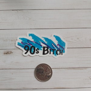 May include: A white sticker with a blue and purple abstract design and the text "90s Bitch" in black. A Canadian quarter is in the foreground.